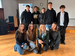 Na zdjęciu widać grupę uczniów wraz z zaproszonym gościem podczas spotkania w szkole. Uczniowie stoją i klęczą wokół kobiety siedzącej z przodu, tworząc pamiątkowe zdjęcie. Wszyscy wyglądają na zadowolonych, a atmosfera jest przyjazna i uroczyst