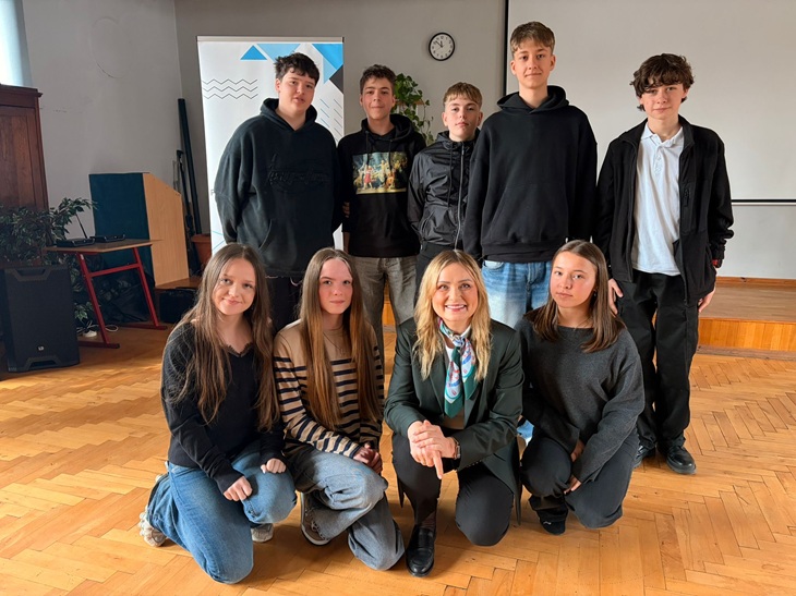 Na zdjęciu widać grupę uczniów wraz z zaproszonym gościem podczas spotkania w szkole. Uczniowie stoją i klęczą wokół kobiety siedzącej z przodu, tworząc pamiątkowe zdjęcie. Wszyscy wyglądają na zadowolonych, a atmosfera jest przyjazna i uroczyst