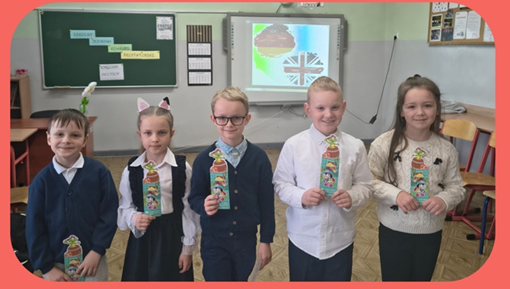 Na zdjęciu widać grupę uczniów stojących w klasie, którzy prezentują zdobyte nagrody lub dyplomy. Za nimi znajduje się tablica oraz ekran z grafiką nawiązującą do języków obcych. Dzieci są elegancko ubrane, uśmiechnięte i wyglądają na dumne ze swoich osiągnięć.