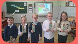 Na zdjęciu widać grupę uczniów stojących w klasie, którzy prezentują zdobyte nagrody lub dyplomy. Za nimi znajduje się tablica oraz ekran z grafiką nawiązującą do języków obcych. Dzieci są elegancko ubrane, uśmiechnięte i wyglądają na dumne ze swoich osiągnięć.