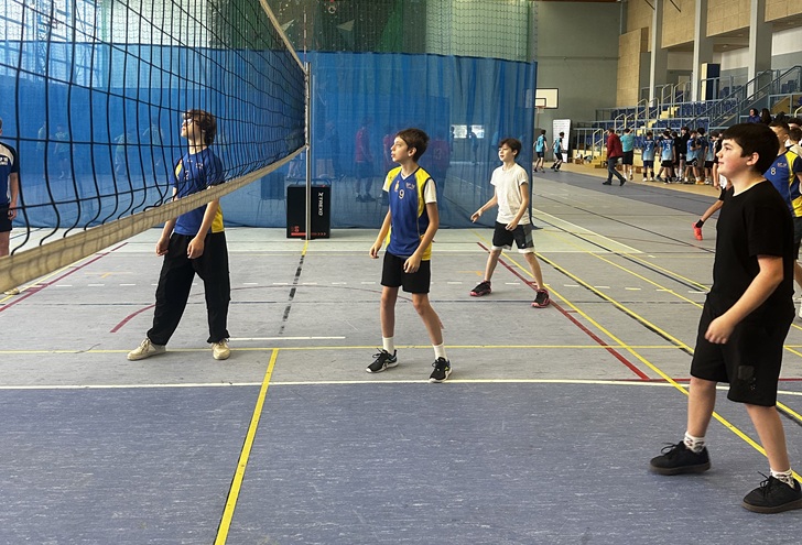 Uczniowie podczas meczu siatkówki na hali sportowej, skupieni i gotowi do odbioru piłki. W tle widać inne drużyny oraz kibiców, co podkreśla sportową atmosferę turnieju