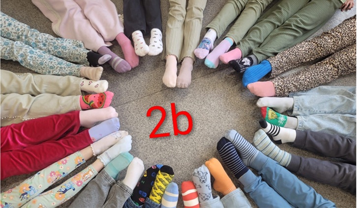 Uczniowie klasy 2b siedzą w kręgu, prezentując kolorowe, niepasujące do siebie skarpetki. Zdjęcie symbolizuje różnorodność, jedność oraz wspólne zaangażowanie w akcję.