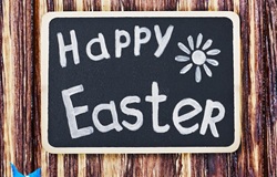 Na obrazku widać tabliczkę z napisem „Happy Easter” napisaną białą kredą na czarnym tle. Tabliczka znajduje się na drewnianym tle, co nadaje całości rustykalny, świąteczny charakter.