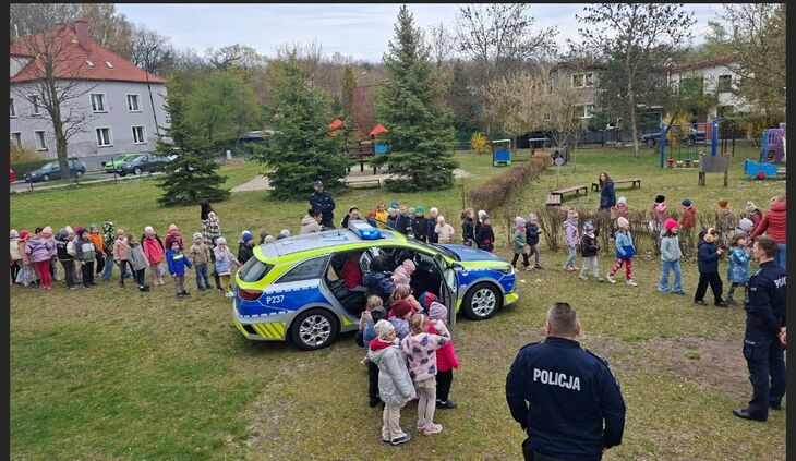 grupa dzieci stojąca w towarzystwie maskotki policyjnej oraz dwójki policjantów w mundurach, w tle radiowóz
