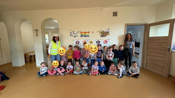 grupa dzieci siedząca w dwóch rzędach wraz z zwychowawcą grupy oraz kobietą w kasku budowlanym i kamizelce odblaskowejpi