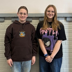 Na zdjęciu widać dwoje uczniów stojących obok siebie na tle ściany z jasnej cegły. Uśmiechają się do zdjęcia i mają swobodny, szkolny strój – jedna osoba ma bluzę z kapturem, druga koszulkę z nadrukiem. Atmosfera jest przyjazna i naturalna.