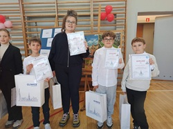 Na zdjęciu widać grupę uczniów stojących w sali gimnastycznej, którzy prezentują zdobyte dyplomy i nagrody. Dzieci trzymają również torby z upominkami, a ich postawa i uśmiechy świadczą o dumie z osiągniętego sukcesu w konkursie.