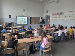 Na zdjęciu widać klasę szkolną podczas zajęć. Uczniowie siedzą przy ławkach i oglądają materiał wyświetlany na tablicy multimedialnej, który dotyczy bezpiecznego korzystania z internetu. Atmosfera jest spokojna, a dzieci są skupione na lekcji.