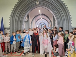 Na zdjęciu widać uczniów uczestniczących w pokazie mody ekologicznej. Dzieci prezentują kolorowe, kreatywne stroje wykonane z różnych materiałów, a wokół nich stoją pozostali uczniowie, obserwując wydarzenie z zainteresowaniem. Atmosfera jest radosna i pełna zaangażowania.