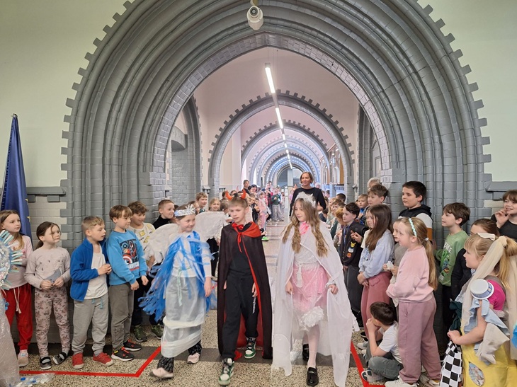 Na zdjęciu widać uczniów uczestniczących w pokazie mody ekologicznej. Dzieci prezentują kolorowe, kreatywne stroje wykonane z różnych materiałów, a wokół nich stoją pozostali uczniowie, obserwując wydarzenie z zainteresowaniem. Atmosfera jest radosna i pełna zaangażowania.