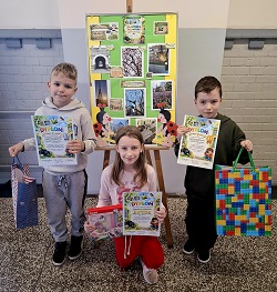 Na zdjęciu widać troje uczniów prezentujących dyplomy oraz nagrody zdobyte w konkursie fotograficznym. Dzieci stoją przed tablicą z pracami konkursowymi, uśmiechnięte i dumne ze swoich osiągnięć.