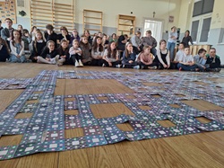 Na zdjęciu widać grupę dzieci siedzących na podłodze w sali gimnastycznej, które oglądają duży, wspólnie ułożony dywan Sierpińskiego 🌀. Przed nimi rozłożone są kolorowe, powtarzalne wzory tworzące fraktal, będący efektem pracy wielu klas.