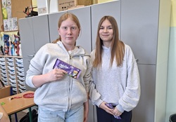 Na zdjęciu widać dwie uczennice stojące w klasie. Dziewczyny trzymają czekolady, które prawdopodobnie otrzymały jako nagrodę za udział w wielkanocnej zabawie. Uśmiechają się i pozują do zdjęcia.