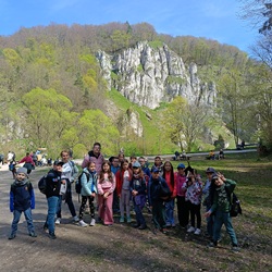 Na zdjęciu widać grupę dzieci wraz z opiekunami podczas wycieczki. Stoją na ścieżce wśród zielonej doliny, a w tle wznoszą się imponujące białe skały wapienne oraz wiosenna roślinność. Uczestnicy wyglądają na zadowolonych i pełnych energii, co dobrze oddaje atmosferę udanej wyprawy.