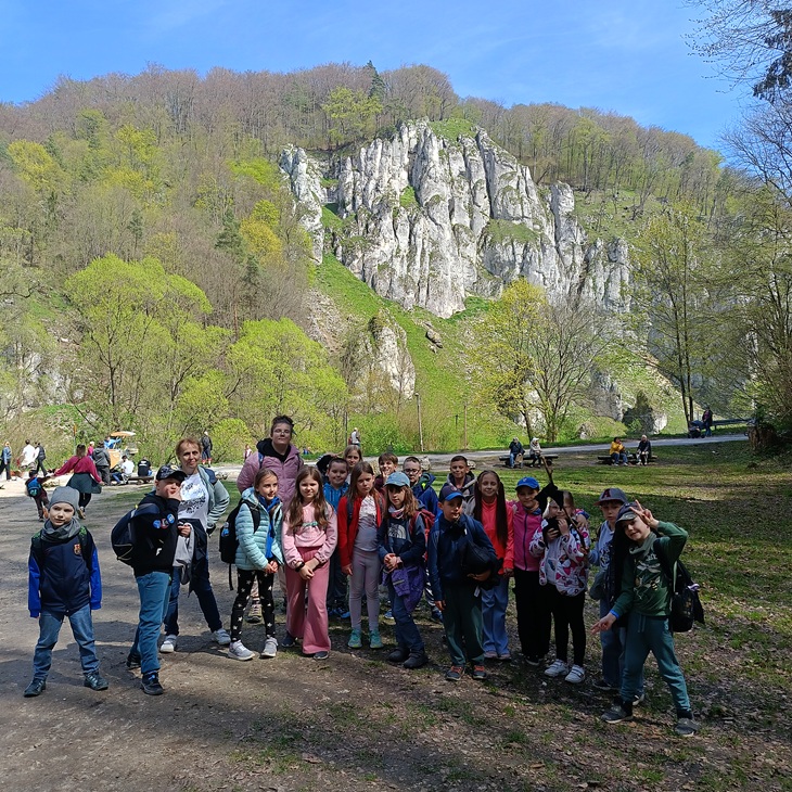Na zdjęciu widać grupę dzieci wraz z opiekunami podczas wycieczki. Stoją na ścieżce wśród zielonej doliny, a w tle wznoszą się imponujące białe skały wapienne oraz wiosenna roślinność. Uczestnicy wyglądają na zadowolonych i pełnych energii, co dobrze oddaje atmosferę udanej wyprawy.
