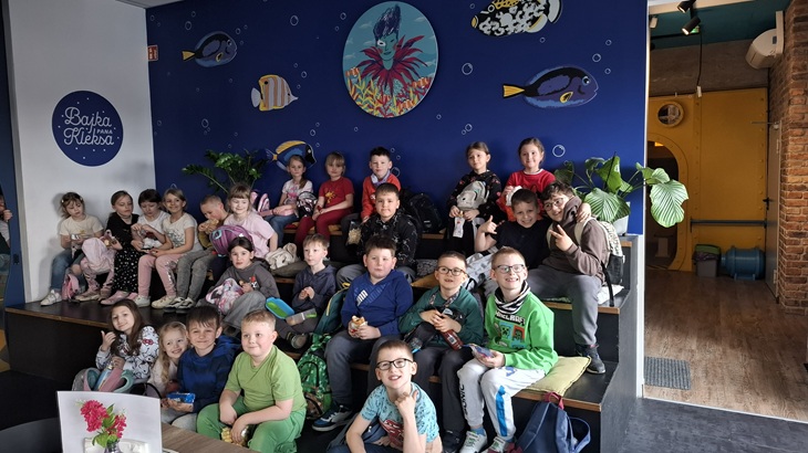 Na zdjęciu widać grupę uczniów klas pierwszych podczas wycieczki do Bajki Pana Kleksa. Dzieci siedzą razem na ławkach, uśmiechnięte i zadowolone, pozując do wspólnej fotografii. W tle znajduje się kolorowa ściana z motywem podwodnego świata, co podkreśla bajkowy i edukacyjny charakter miejsca.