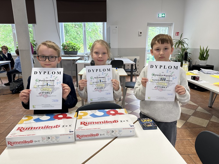 Na zdjęciu widać troje dzieci stojących w sali lekcyjnej, które z dumą prezentują swoje dyplomy z konkursu matematycznego. Przed nimi leżą nagrody – gry planszowe, m.in. Rummikub i UNO. Uczniowie wyglądają na zadowolonych i dumnych ze swoich osiągnięć.