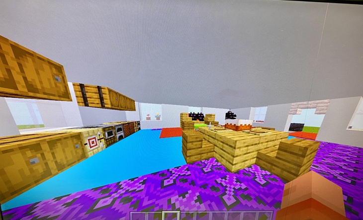 Na obrazie widać wnętrze stworzone w grze Minecraft – jasne, kolorowe pomieszczenie przypominające kuchnię lub jadalnię. Znajdują się tu drewniane blaty z ustawionymi tortami, szafki, piekarniki oraz dekoracje. Podłoga jest wielobarwna, a całość wygląda na starannie zaprojektowaną i kreatywnie urządzoną przestrzeń.