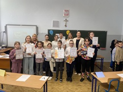 Na zdjęciu widać grupę uczniów klas trzecich wraz z nauczycielkami w sali lekcyjnej. Dzieci trzymają dyplomy i nagrody, pozując po zakończonym konkursie „Mistrz savoir-vivre”. Wszyscy wyglądają na zadowolonych i dumni z osiągnięć.