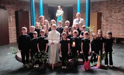 Na zdjęciu widać grupę dzieci z klasy 2c wraz z nauczycielkami podczas występu o tematyce wielkanocnej. Uczniowie stoją w kościele, ubrani na czarno, a jedno z dzieci ma białą szatę i trzyma drewniany krzyż. W tle znajduje się figura Matki Bożej z Dzieciątkiem. Dzieci trzymają także symbole związane z przedstawieniem, co podkreśla uroczysty i refleksyjny charakter wydarzenia.