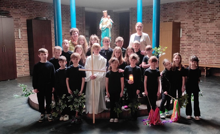 Na zdjęciu widać grupę dzieci z klasy 2c wraz z nauczycielkami podczas występu o tematyce wielkanocnej. Uczniowie stoją w kościele, ubrani na czarno, a jedno z dzieci ma białą szatę i trzyma drewniany krzyż. W tle znajduje się figura Matki Bożej z Dzieciątkiem. Dzieci trzymają także symbole związane z przedstawieniem, co podkreśla uroczysty i refleksyjny charakter wydarzenia.