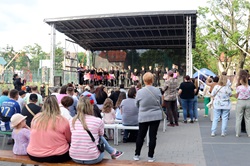Na zdjęciu widać rodzinny festyn odbywający się na świeżym powietrzu. W centrum znajduje się scena z zadaszeniem, na której występują dzieci prezentujące program artystyczny. Przed sceną zgromadzili się uczestnicy wydarzenia – rodzice, dzieci oraz mieszkańcy, którzy oglądają występy i wspólnie spędzają czas. Część osób siedzi na ławkach, inni stoją i obserwują występy. Całości towarzyszy pogodna, serdeczna atmosfera rodzinnego spotkania.