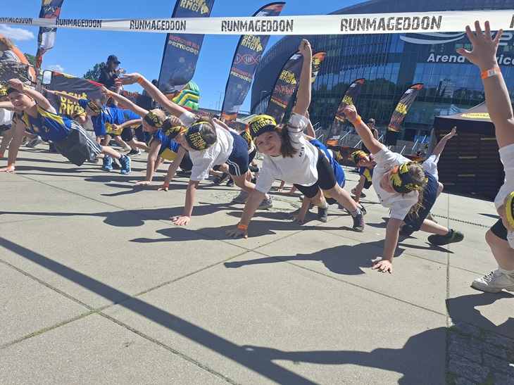 Na zdjęciu widać grupę dzieci biorących udział w wydarzeniu Runmageddon Kids. Uczestnicy, ubrani w stroje sportowe i charakterystyczne opaski na głowach, wykonują ćwiczenie ruchowe pod specjalną bramą z napisem „Runmageddon”. Dzieci poruszają się w podporze, unosząc jedną rękę ku górze, co wymaga dobrej koordynacji i równowagi.  W tle znajdują się flagi wydarzenia oraz nowoczesny budynek hali widowiskowo-sportowej. Słoneczna pogoda i zaangażowanie uczestników podkreślają sportowy charakter imprezy. Zdjęcie oddaje atmosferę aktywności, współpracy i radości z podejmowania nowych wyzwań.