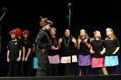 Na zdjęciu widać grupę dzieci występujących na scenie podczas przedstawienia teatralnego. Uczniowie ubrani są w czarne stroje sceniczne, a część dziewczynek ma kolorowe spódniczki i ozdoby we włosach. Po lewej stronie stoją chłopcy w czerwonych hełmach strażackich, co sugeruje, że spektakl dotyczy pracy strażaków lub akcji ratunkowej. W centralnej części sceny znajduje się chłopiec w kostiumie przypominającym zwierzę lub bajkową postać, który odgrywa główną rolę i zwraca uwagę swoją postawą oraz strojem. Pozostali uczestnicy śpiewają lub recytują swoje kwestie, gestykulując i angażując się w występ. Na scenie widoczne są mikrofony oraz ciemne tło kurtyny teatralnej, które podkreślają charakter wydarzenia artystycznego. Zdjęcie oddaje atmosferę dziecięcego spektaklu pełnego emocji, zaangażowania i współpracy.
