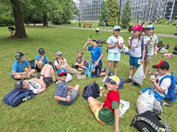 Na zdjęciu widać grupę dzieci odpoczywających na trawie w parku. Większość siedzi lub leży na trawniku, a wiele z nich trzyma lody w wafelkach i je zajada. Dzieci mają na sobie letnie ubrania – koszulki, krótkie spodenki oraz czapki z daszkiem chroniące przed słońcem. Wokół nich leżą plecaki, torby i rower.  W tle znajdują się drzewa, ławki parkowe oraz ścieżka spacerowa. Po prawej stronie widać dużą drewnianą konstrukcję przypominającą fragment kolejki górskiej lub inną dużą atrakcję rekreacyjną. Pogoda wydaje się ciepła i przyjemna, a cała scena sprawia wrażenie wycieczki lub wspólnego wypoczynku grupy dzieci podczas letniego dnia.