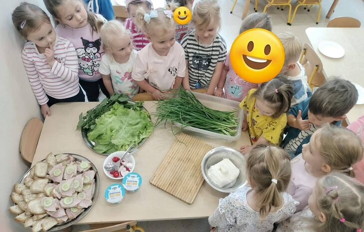 grupa dzieci stojąca w sali przedszkolnej przy stole, na stole szczypiorek, twaróg i pieczywo gotowe do przygotowania kanapek
