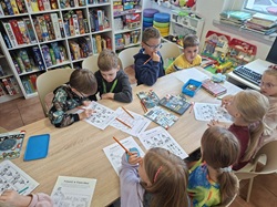 Na zdjęciu widać dzieci podczas warsztatów w bibliotece. Uczniowie siedzą przy wspólnym stole i rozwiązują przygotowane zagadki oraz łamigłówki detektywistyczne. Wokół nich znajdują się książki, gry planszowe i materiały edukacyjne. Dzieci pracują w skupieniu, samodzielnie oraz w grupach, rozwijając logiczne myślenie i współpracę.