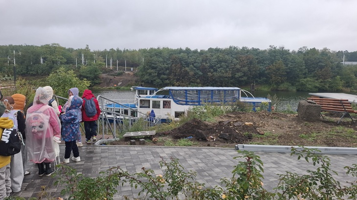 Na zdjęciu widać grupę uczniów przygotowujących się do rejsu po Kanale Gliwickim. Uczestnicy, ubrani w przeciwdeszczowe kurtki i peleryny, schodzą w kierunku nabrzeża, gdzie czeka na nich statek.  W tle rozciąga się spokojna tafla wody oraz otaczająca kanał zieleń. Mimo pochmurnej i deszczowej pogody atmosfera wydaje się pogodna i pełna oczekiwania na wspólną przygodę.  Zdjęcie przedstawia początek wodnej wyprawy i edukacyjnej przygody uczniów.