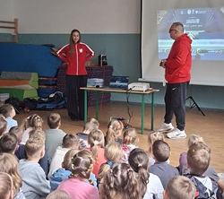 Na zdjęciu widać spotkanie uczniów z przedstawicielami świata sportu. W sali gimnastycznej przed zgromadzonymi dziećmi stoi młoda zawodniczka w stroju sportowym oraz mężczyzna prowadzący spotkanie. W tle znajduje się ekran z prezentacją multimedialną, a obok przygotowany jest sprzęt sportowy. Uczniowie z zainteresowaniem obserwują rozmowę i prezentację. Zdjęcie pokazuje edukacyjne i inspirujące spotkanie, podczas którego dzieci miały okazję poznać bliżej sport oraz życie młodej zawodniczki.