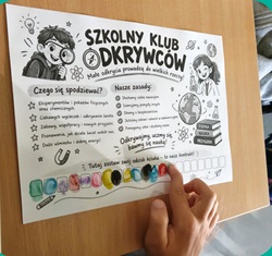 Na zdjęciu widać kartę informacyjną Szkolnego Klubu Odkrywców. Karta znajduje się na ławce, a dziecko wskazuje palcem jeden z jej elementów. Na plakacie przedstawiono kolorowe, dziecięce ilustracje związane z nauką i odkrywaniem świata, m.in. lupę, żarówkę, probówki oraz postacie młodych odkrywców. Znajdują się na nim również informacje dotyczące działalności Klubu, między innymi tego, czego uczniowie będą poszukiwać i jakie zasady będą obowiązywać podczas zajęć. Zdjęcie pokazuje ciekawą i angażującą formę zaprezentowania dzieciom zajęć przyrodniczych, zachęcającą je do eksperymentowania, zadawania pytań i odkrywania świata.